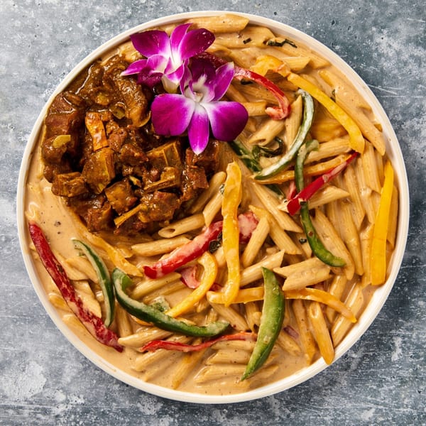 Curried goat and penne pasta with bell peppers in a creamy sauce, garnished with purple orchids.