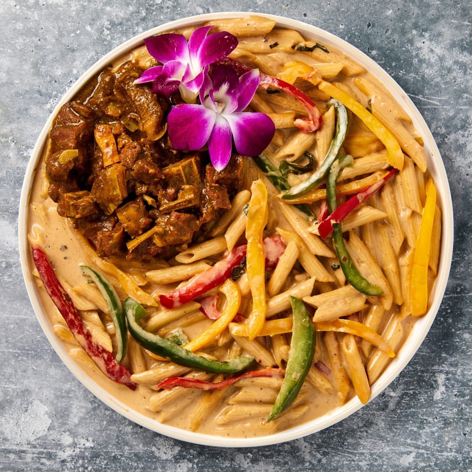 Curried goat and penne pasta with bell peppers in a creamy sauce, garnished with purple orchids.