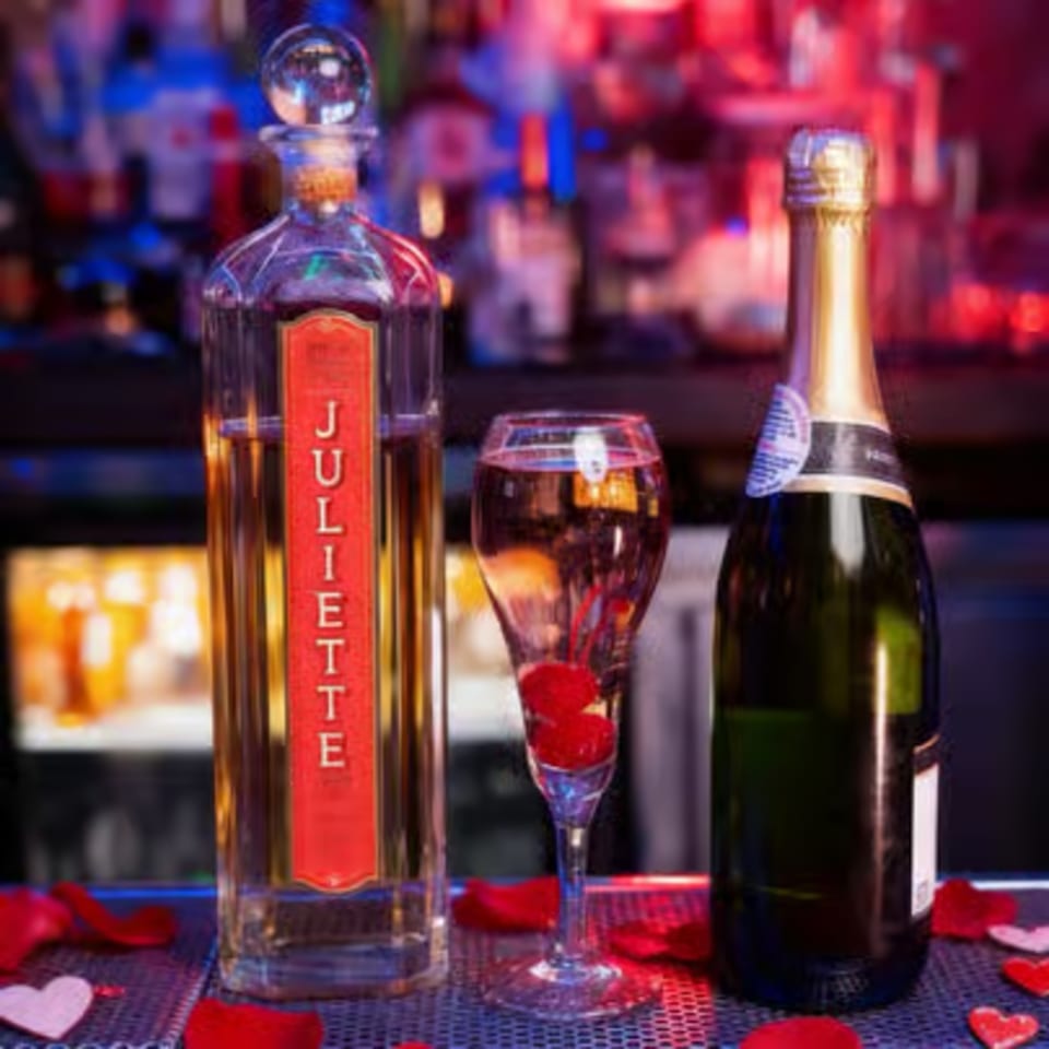 A Juliette liquor bottle, champagne flute with cherries, and champagne bottle on a bar with rose petals.