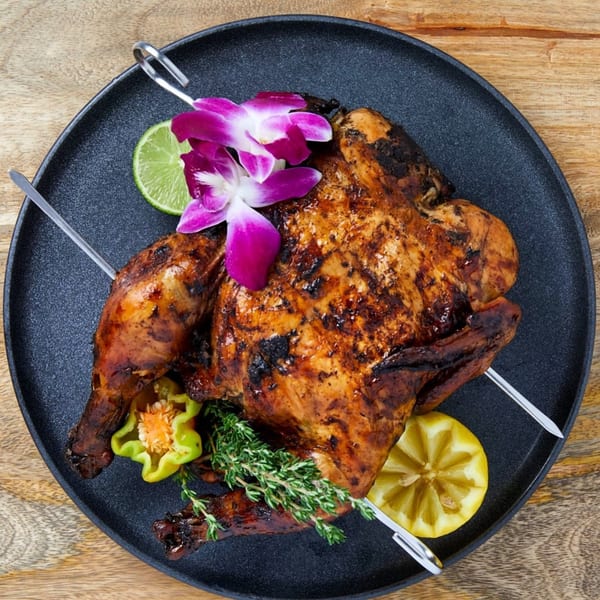 Whole roasted chicken on a black plate, garnished with orchids, lime, lemon, and herbs.