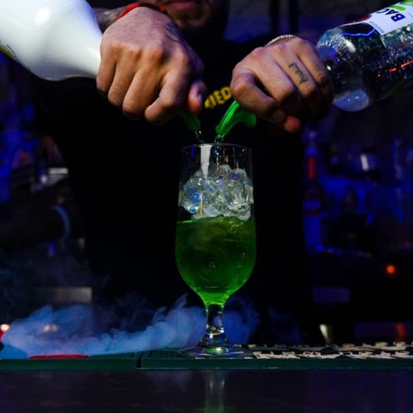 Bartender pouring two liquids into a glass with ice and green liquid, surrounded by smoke.