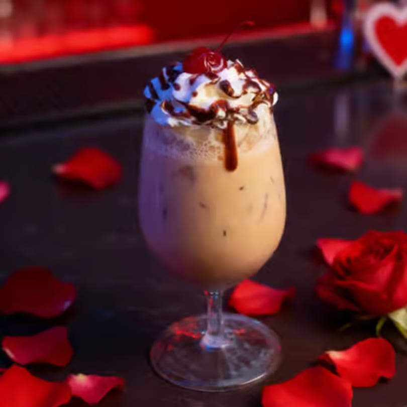 Iced coffee with whipped cream, chocolate drizzle, and a cherry, surrounded by red rose petals.