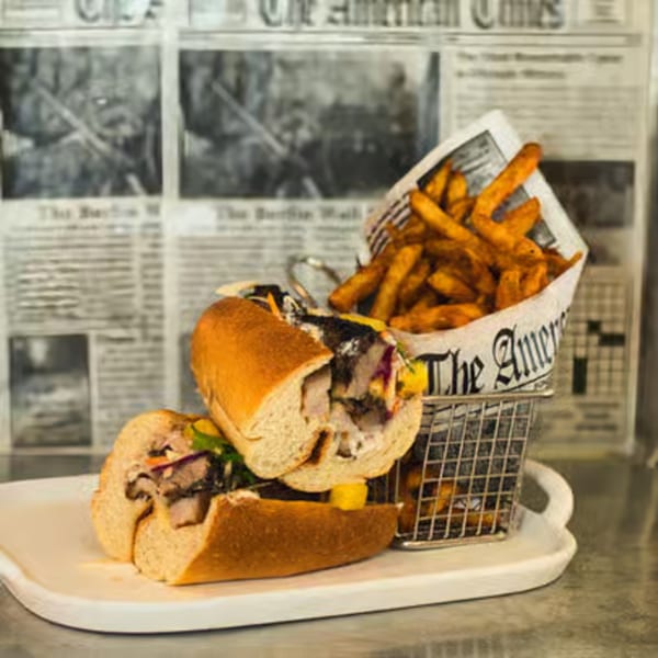 A pork sandwich with colorful toppings and a side of fries in a metal basket on a white tray.