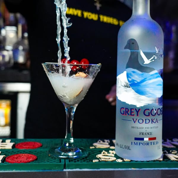 Grey Goose vodka bottle next to a martini glass with liquid pouring into it, garnished with cherries.