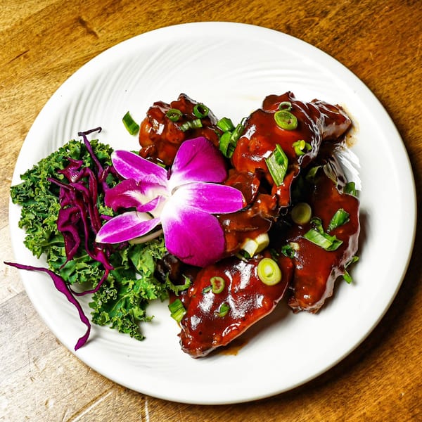 Chicken wings coated in a dark sauce, garnished with green onions, kale, red cabbage, and a purple orchid.